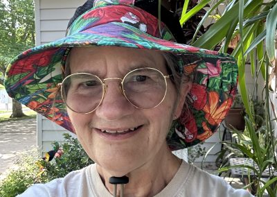 Becky Rawls-Riley wearing a handcrafted boonie hat among garden plants, reflecting her artisan headwear design work.